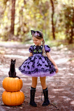 Load image into Gallery viewer, A purple and black bat and cat themed twirl dress stands on a forest path next to a black cat sitting on stacked pumpkins. The dress features a black cat face graphic on the bodice and black bat silhouettes on the skirt.
